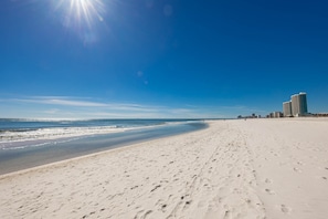 A wide sandy beach with footprints leading to the water, a clear blue sky, and a distant cityscape with high-rise buildings. - Phoenix Viii 8106 3 Bedroom Condo (Orange Beach)