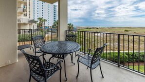 A balcony with a round table and chairs, overlooking a beach and a hotel building. - Phoenix Vii 7117 3 Bedroom Condo (Orange Beach)