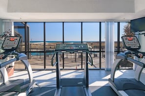 Ein Fitnessraum mit Laufbändern und einem Ergometer, große Fenster mit Blick auf den Strand.