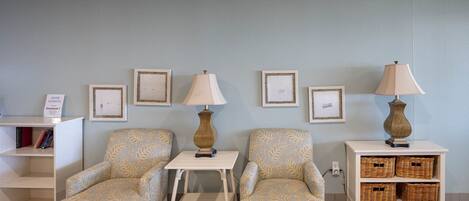 A waiting area with two patterned armchairs, a small table, and a side table with a lamp. There are framed pictures on the wall and a bookshelf with books.