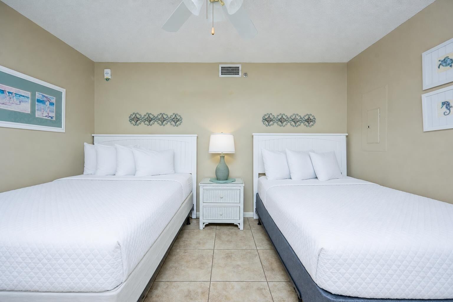 A hotel room with two beds, a nightstand with a lamp, decorative wall art, and a ceiling fan.