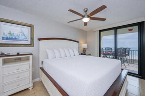 Ein Hotelzimmer mit einem Bett, einem Nachttisch, einer Lampe, einem Deckenventilator, einem gerahmten Bild und Blick auf den Strand durch große Fenster.