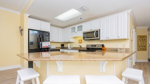 A kitchen with white cabinets, a granite countertop, a stainless steel refrigerator, a microwave, and two white bar stools. - Phoenix Vii 71205 3 Bedroom Condo (Orange Beach)