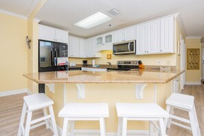 A kitchen with white cabinets, a granite countertop, a stainless steel refrigerator, a microwave, and two white bar stools. - Phoenix Vii 71205 3 Bedroom Condo (Orange Beach)