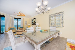 A dining area with a glass table, white chairs, a chandelier, and a large window with curtains. - Phoenix Vii 71205 3 Bedroom Condo (Orange Beach)