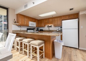 A kitchen with wooden cabinets, a white refrigerator, a microwave, and a bar stool area. - Phoenix Vii 71001 3 Bedroom Condo (Orange Beach)