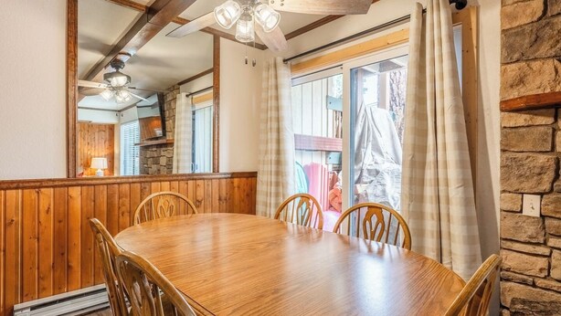 Una taula de dinar de fusta amb cadires, un ventilador de sostre, parets de pedra i una finestra amb cortines.