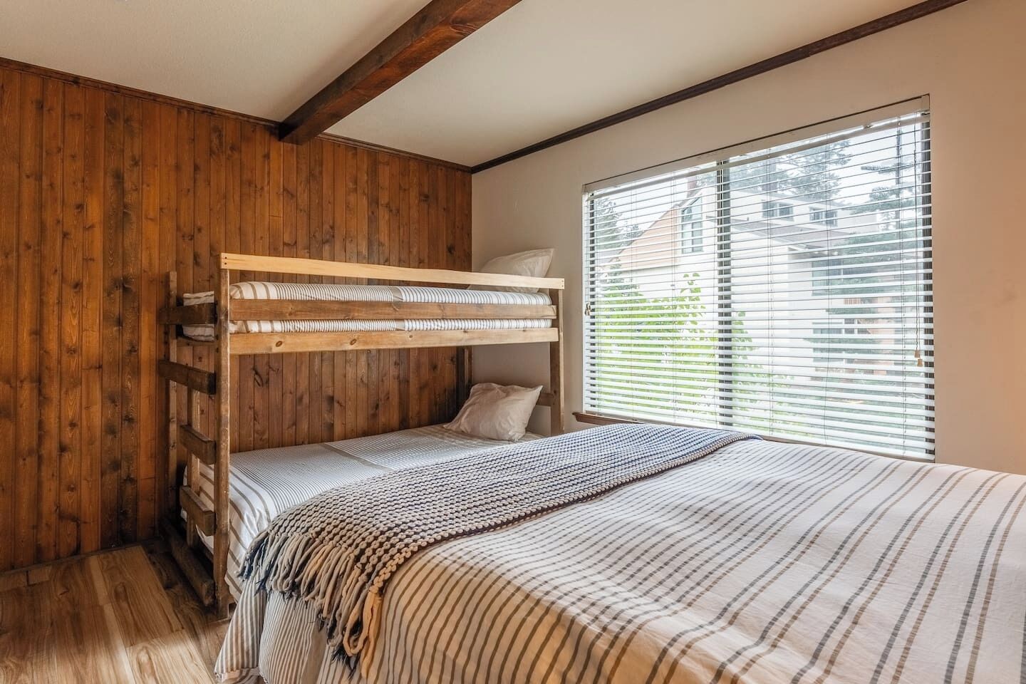 Ein Stockbett mit Holz-Kopfteil, einem Fenster mit Jalousien und einer gestreiften Bettdecke.