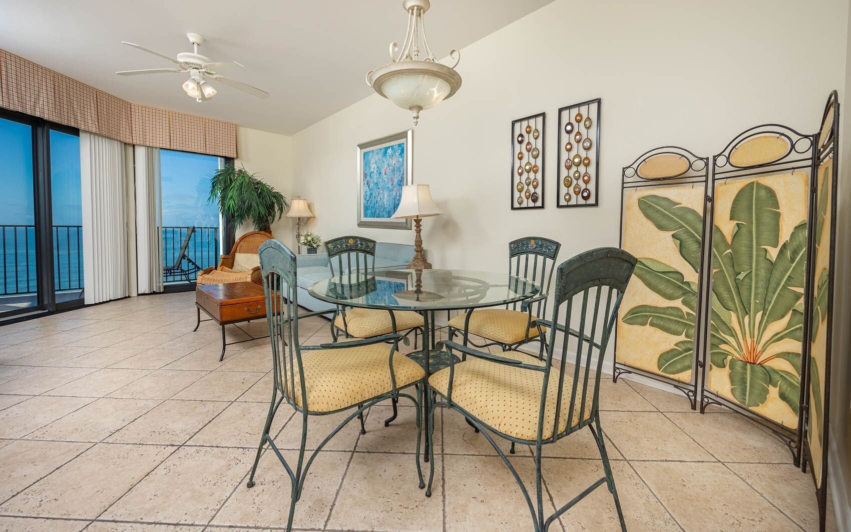 A dining area with a glass table, metal chairs, a potted plant, and artwork on the walls.