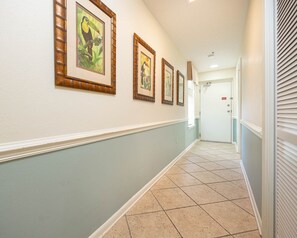 A hallway with framed pictures on the wall, a door at the end, and a tiled floor. - Phoenix Vii 71213 2 Bedroom Condo (Orange Beach)