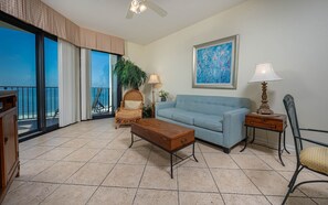 A living room with a blue sofa, a wooden coffee table, a wicker chair, a lamp, and a painting on the wall. - Phoenix Vii 71213 2 Bedroom Condo (Orange Beach)