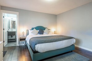 Ein Schlafzimmer mit einem Bett, Nachttischen, einem Schminktisch und einem Spiegel. Ein Badezimmer ist durch eine offene Tür sichtbar.