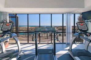 Ein Fitnessraum mit Laufbändern und einem Ergometer, große Fenster mit Blick auf den Strand.