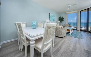 Uma sala de jantar com uma mesa e cadeiras brancas, um ventilador de teto e vista para o oceano através de amplas janelas.