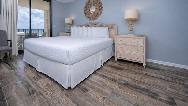 A bedroom with a large bed, a nightstand with lamps, a decorative mirror, and a view through a sliding door. - Phoenix Viii 8502 2 Bedroom Condo (Orange Beach)