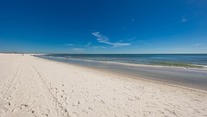 A wide sandy beach with footprints leading to the water, a clear blue sky, and a distant pier. - Phoenix Viii 8502 2 Bedroom Condo (Orange Beach)
