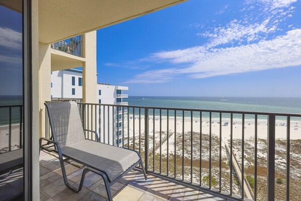 A balcony with a chair, overlooking a beach with a clear sky and a building in the background. - Phoenix Viii 81104 3 Bedroom Condo (Orange Beach)