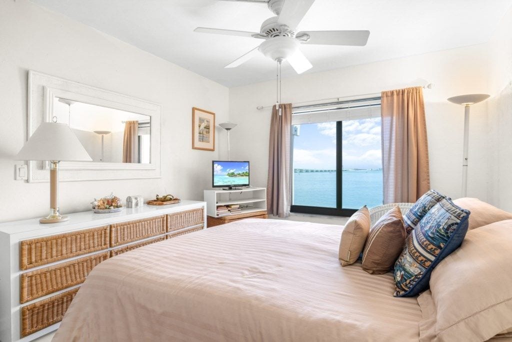A hotel room with a bed, a television, a ceiling fan, a window with curtains, and a view of the sea.