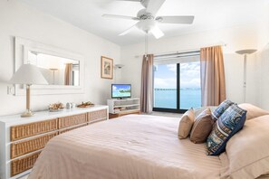 Ein Hotelzimmer mit Bett, Fernseher, Deckenventilator, einem Fenster mit Vorhängen und Meerblick.