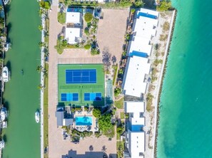 Ett flygbildsbild av en fastighet vid kusten med en tennisbana, en swimmingpool och en strandpromenad.