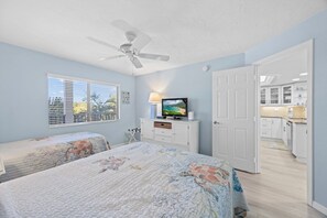 A bedroom with two beds, a ceiling fan, a TV, and a view of the kitchen. - Pointe Santo Unit C22 2 Bedroom Condo (Sanibel)