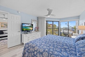 Una camera da letto con un letto grande, una TV, un balcone con vista sulle palme e una ventola a soffitto.