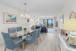 A modern living room with a glass dining table, blue chairs, a sofa, and a balcony with a view. - Pointe Santo Unit C22 2 Bedroom Condo (Sanibel)