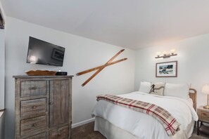 Ein Schlafzimmer mit einem Bett, einem Fernseher auf einer Kommode, einem Wandbild und einem Nachttisch mit einer Lampe.