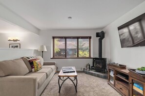 Ein Wohnzimmer mit einer Couch, einem Couchtisch, einem Kamin, einem an der Wand befestigten Fernseher und einem Fenster mit Blick auf Bäume.