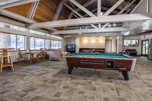 A pool table in a room with a high ceiling and exposed beams, a seating area with chairs, and a television. - Stunning Sunlit & Spacious-1BR/2BTH+Loft (Mammoth Lakes)