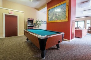 A pool table in a room with a carpeted floor, a door with an exit sign, a framed painting on the wall, and a red couch. - Adventure Basecamp! 1BR + Loft Close to Village (Mammoth Lakes)