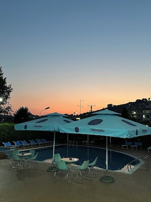 Zona de piscina con sillones de descanso y sombrillas, piscina y un atardecer de fondo.
