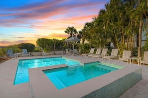 Ein rechteckiger Pool mit zwei Bereichen, umgeben von Liegestühlen und Palmen, vor einem Sonnenuntergangshimmel.