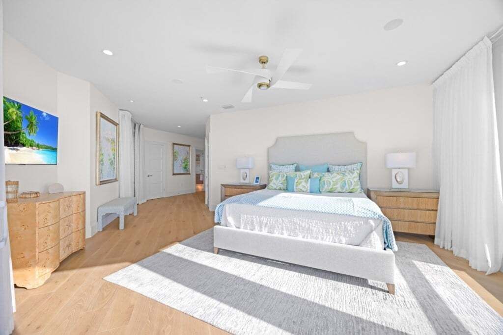 A spacious bedroom with a large bed, a ceiling fan, and a view of a beach through the window.