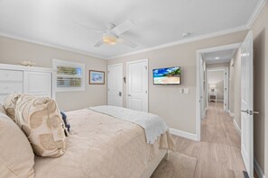 A bedroom with a bed, a ceiling fan, a TV, and a hallway leading to another room. - Seashells Unit 30 - Driftwood Dreams 2 Bedroom Condo (Sanibel)