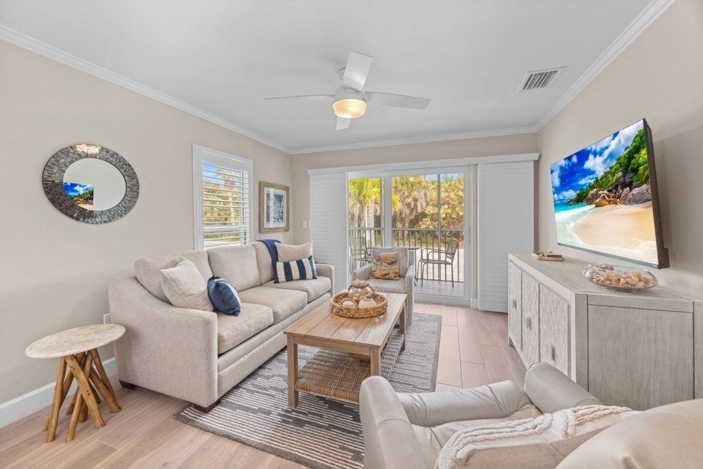 A living room with a sofa, coffee table, and a television. There is a balcony with a view of the outdoors.