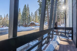 Une terrasse en bois, le sol recouvert de neige, une balustrade en bois et une vue sur un bâtiment dont le toit est également enneigé.