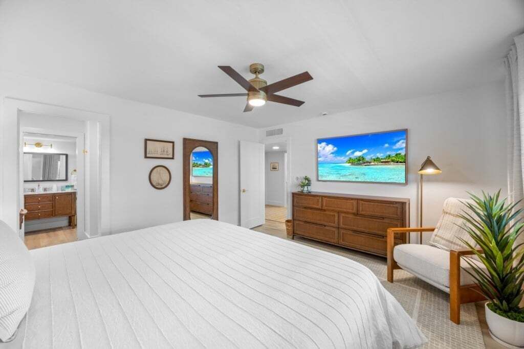 Un dormitorio con cama, ventilador de techo, cómoda de madera, una silla, televisor y puerta que da a otra habitación.
