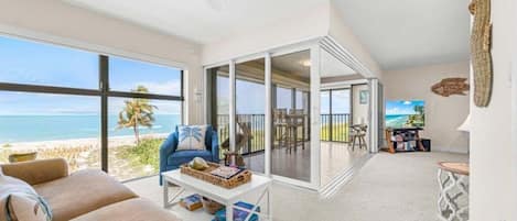 A spacious living area with a large window overlooking a beach, a comfortable seating area, and a view of the ocean.