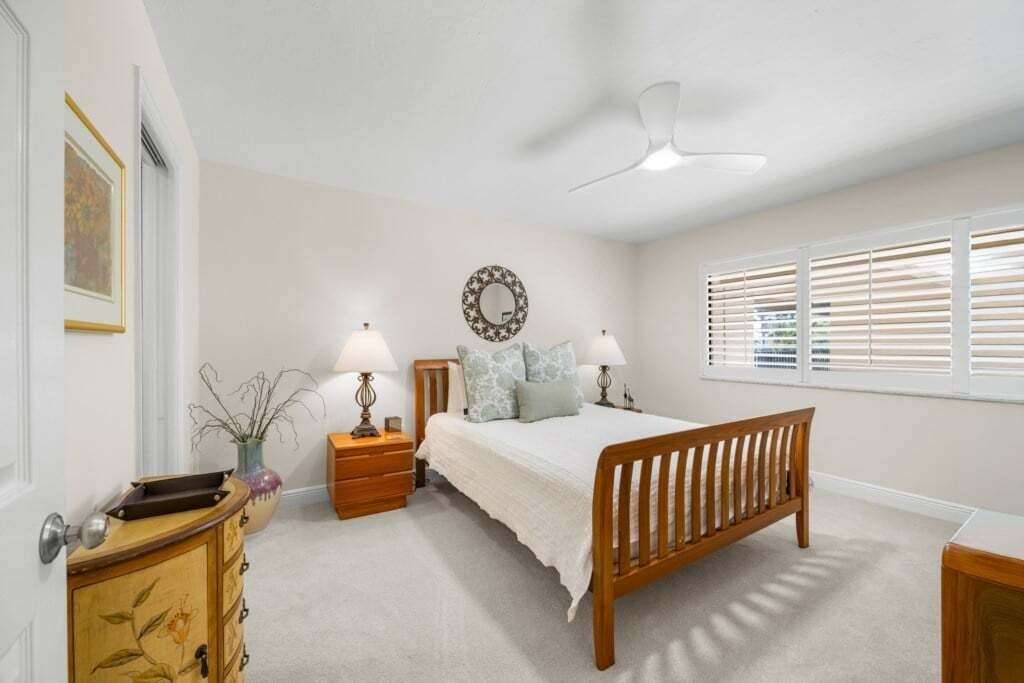 A bedroom with a bed, bedside tables, a ceiling fan, a window with blinds, and a wall-mounted mirror.