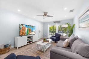 A modern living room with a flat-screen TV, a ceiling fan, a sofa, a coffee table, and a view of the beach through a glass door. - Seashells Unit 32 2 Bedroom Condo (Sanibel)