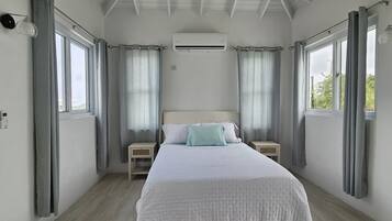 A bedroom with a bed, two bedside tables, an air conditioning unit, and large windows with curtains.