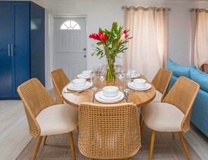 A dining area with a round table set for six, surrounded by wicker chairs, a vase with flowers, and a blue cabinet in the background. - Kaz Olivia 3 Bedroom Home (Gros Islet)