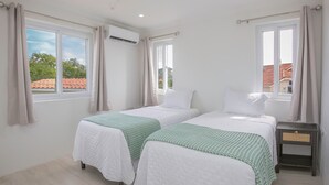 Quarto com duas camas, cada uma com colcha branca e manta verde. Há janelas com cortinas, um ar condicionado e uma mesa de cabeceira com gaveta.