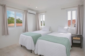 Ein Zimmer mit zwei Betten, jeweils mit weißer Bettdecke und grüner Tagesdecke. Es gibt Fenster mit Vorhängen, eine Klimaanlage und einen Nachttisch mit Schublade.