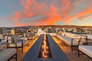 A rooftop terrace with a long fire feature, seating areas, and a view of a residential area with buildings and mountains in the background. - Paradise Palace 8 Bedroom Home (St. George)