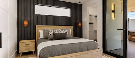 Un dormitorio moderno con cama, mesitas de noche, un espejo grande y puerta corrediza de vidrio que da a un balcón.