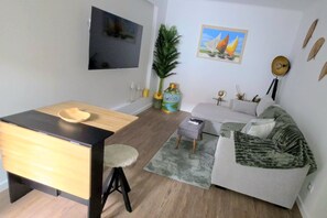 Un espacio habitable compacto con un sofá, una mesita, un televisor montado en la pared y una planta decorativa.