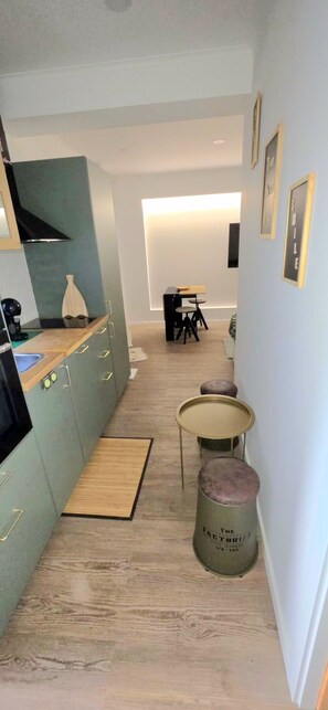A narrow hallway with a kitchen on one side, a dining area with a table and chairs, and a small stool with a label.