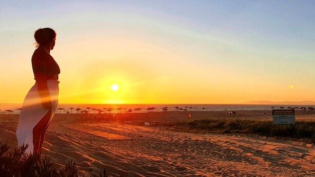 A beach scene with a person standing on the sand, sun setting in the background, and beach umbrellas in the distance.